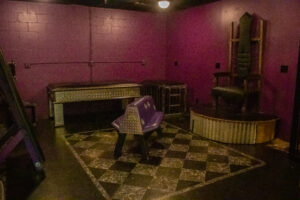 Image of a room with a bordered black and silver tile floor, purple walls, and purple kink furniture